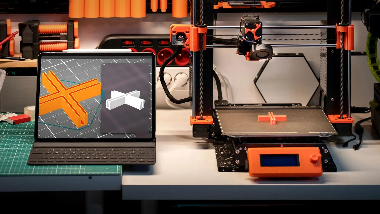 Tablet showing a 3D model of an orange cross-shaped part next to a 3D printer printing the same part