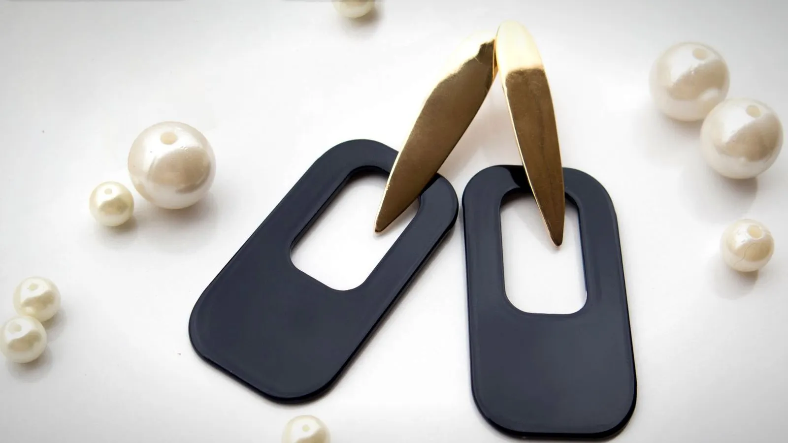 Black geometric statement earrings with gold-colored accents next to pearls on a white surface