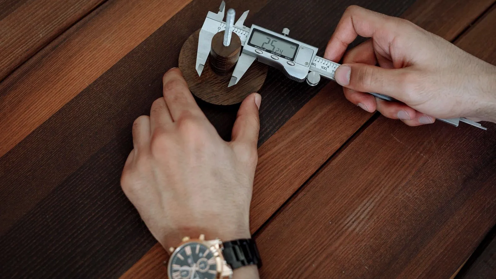 Hands measuring a small turned wooden part with a digital caliper on a wooden table