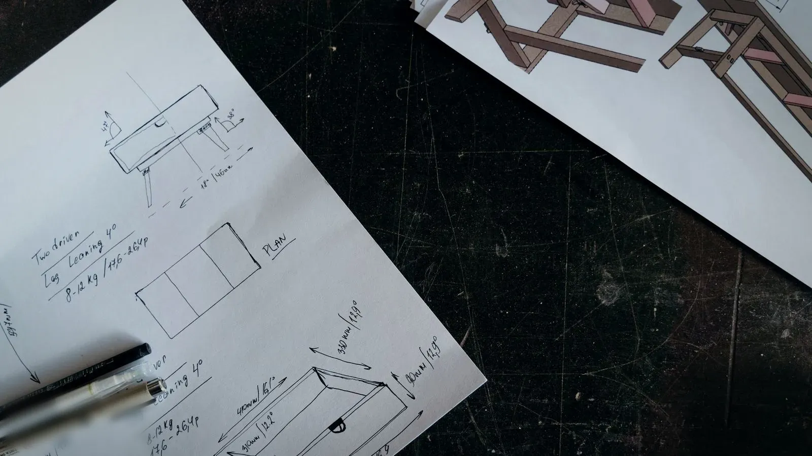 Hand-drawn sketches of a box-shaped part with dimension lines, on paper next to two pens