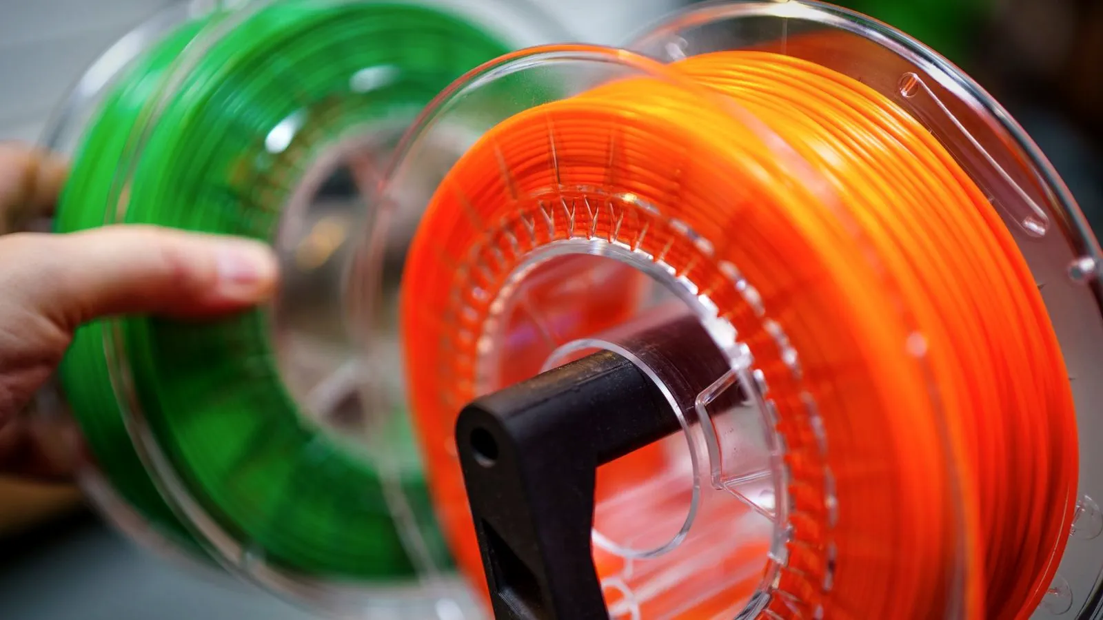 Hand placing an orange filament spool on a spool holder next to a green spool