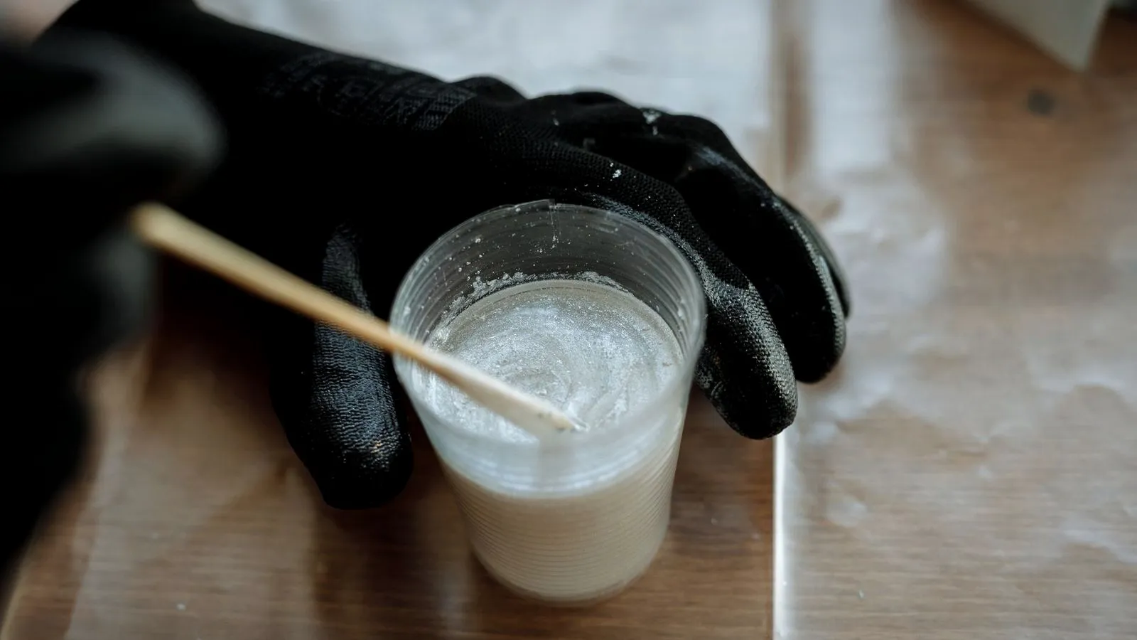 Gloved hands stirring liquid resin in a plastic cup with a wooden stick