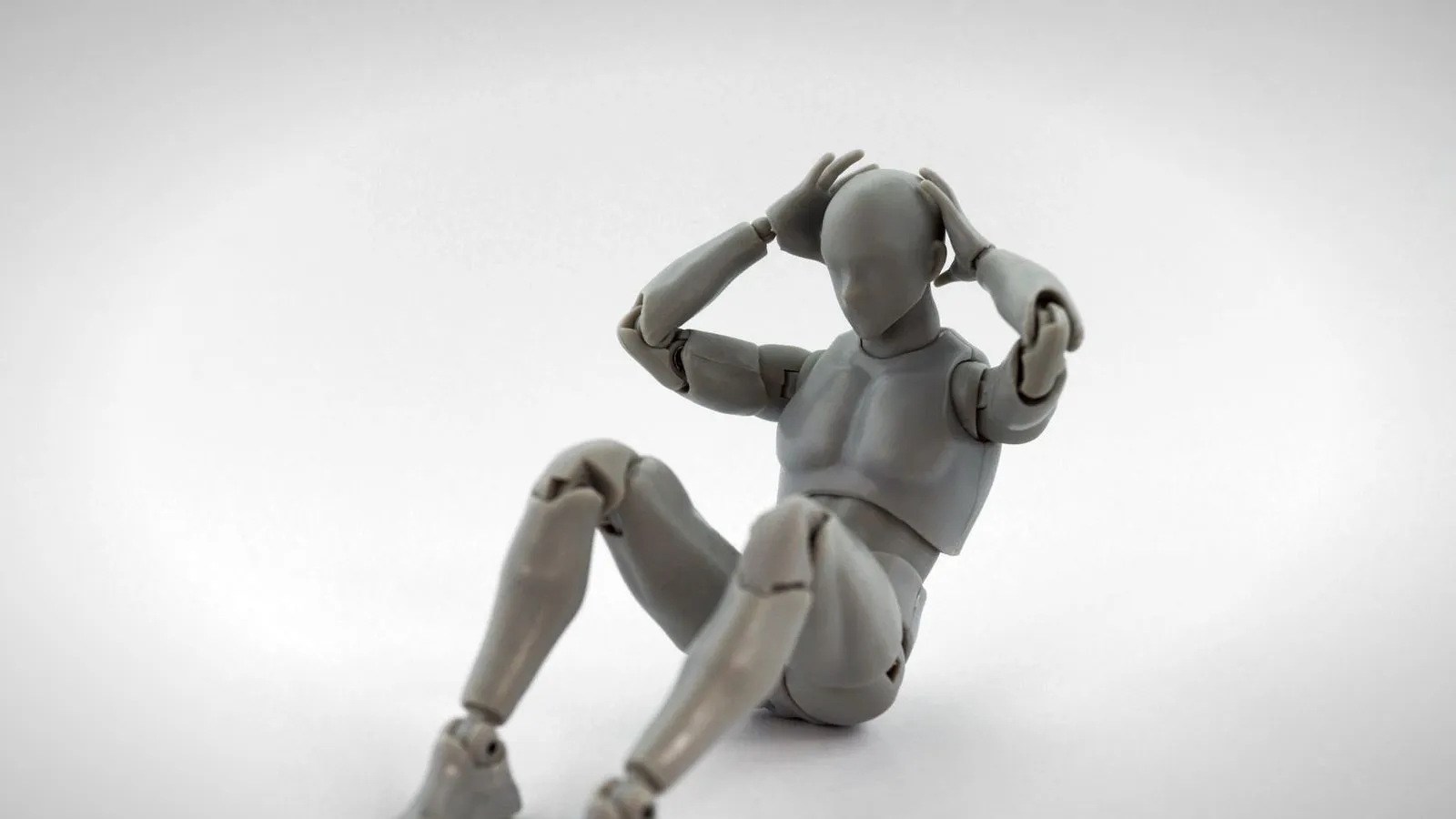 Gray plastic articulated figure with jointed limbs, sitting with its hands on its head