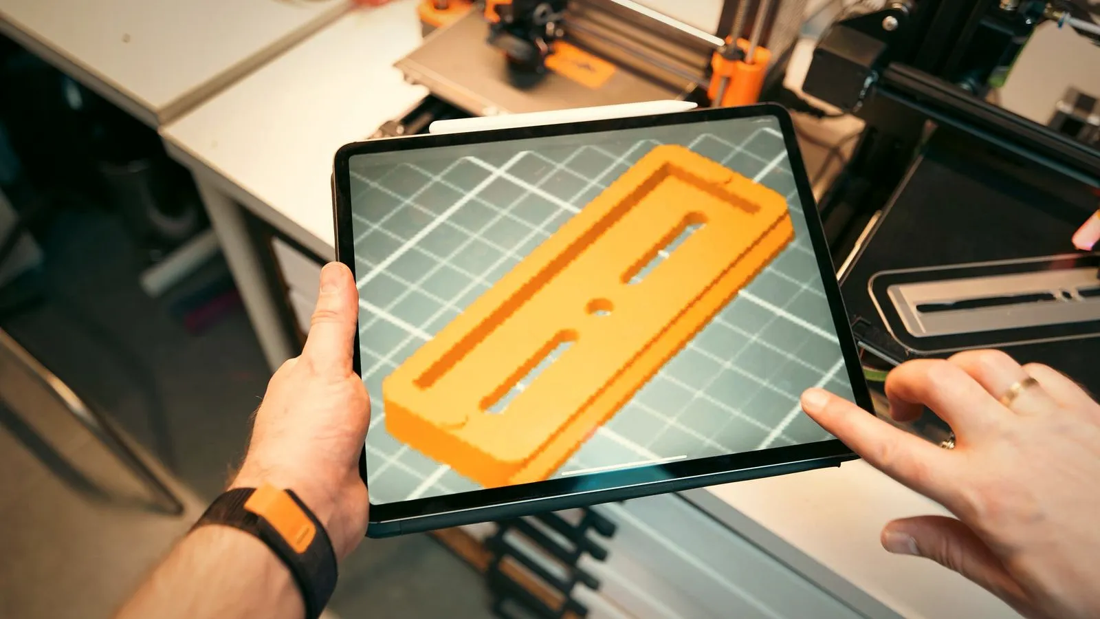 Hands holding a tablet that shows an orange 3D printed part model on a grid, with 3D printers in the background