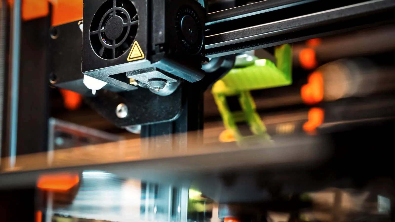 3D printer hotend with its cooling fan above the bed, a green printed part in the background
