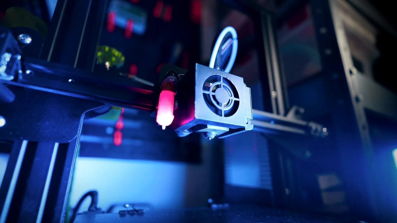 Print head with a metal fan shroud over the hotend and nozzle, a bed probe glowing red beside it, lit in blue