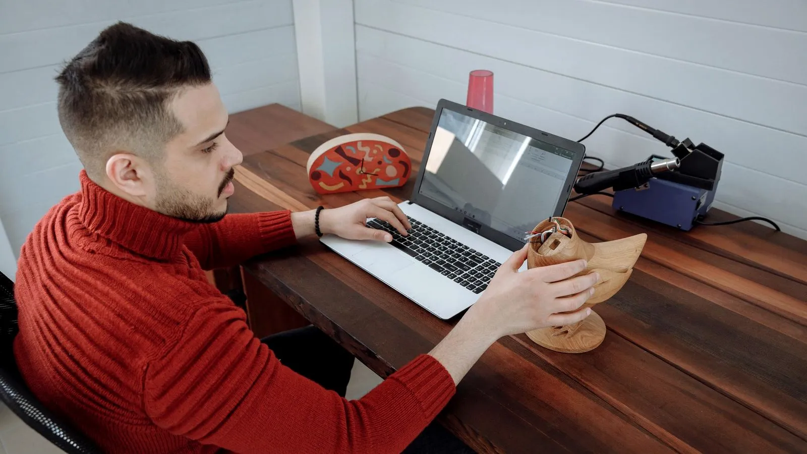 A man at a laptop on a wooden table holding a wood-look bird figure, with a soldering station beside him