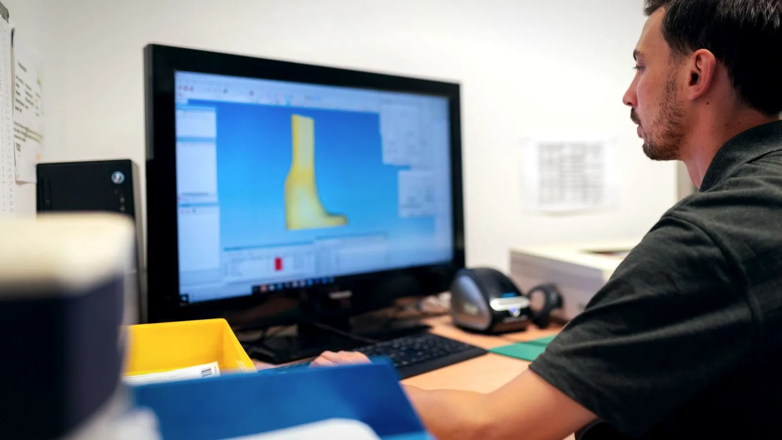 A man at a desk monitor showing a yellow 3D model on screen