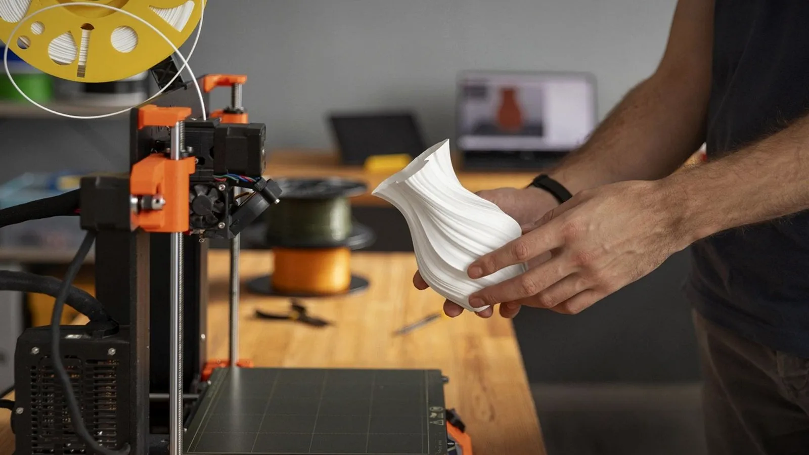 Man holding a white 3D printed vase next to an open-frame 3D printer on a workbench