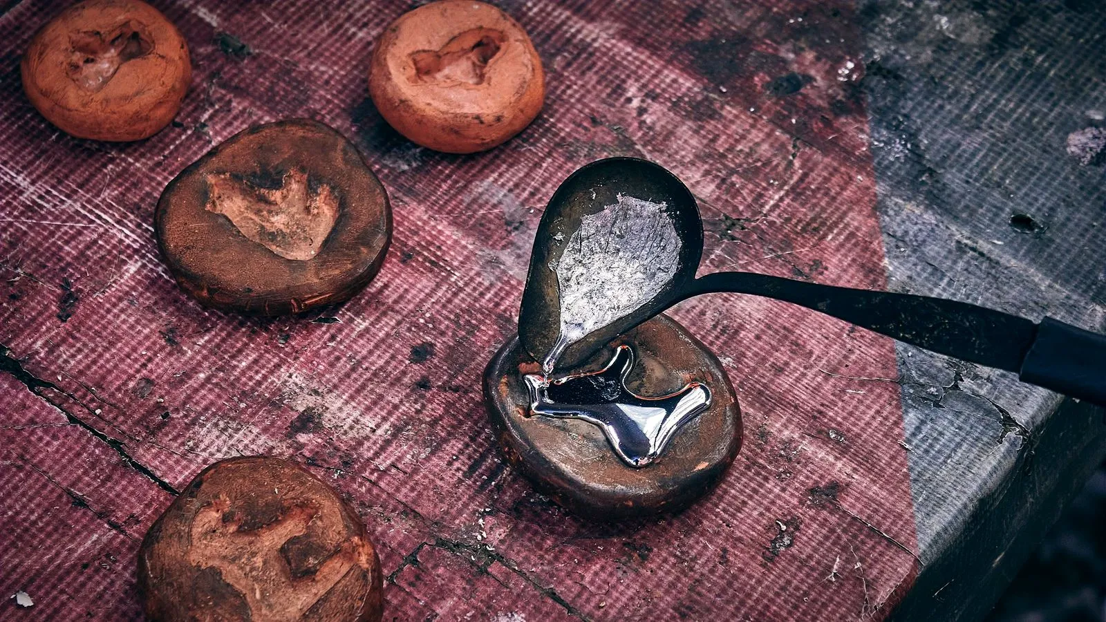 Molten metal poured from a small ladle into a clay mold, with more empty molds on the workbench