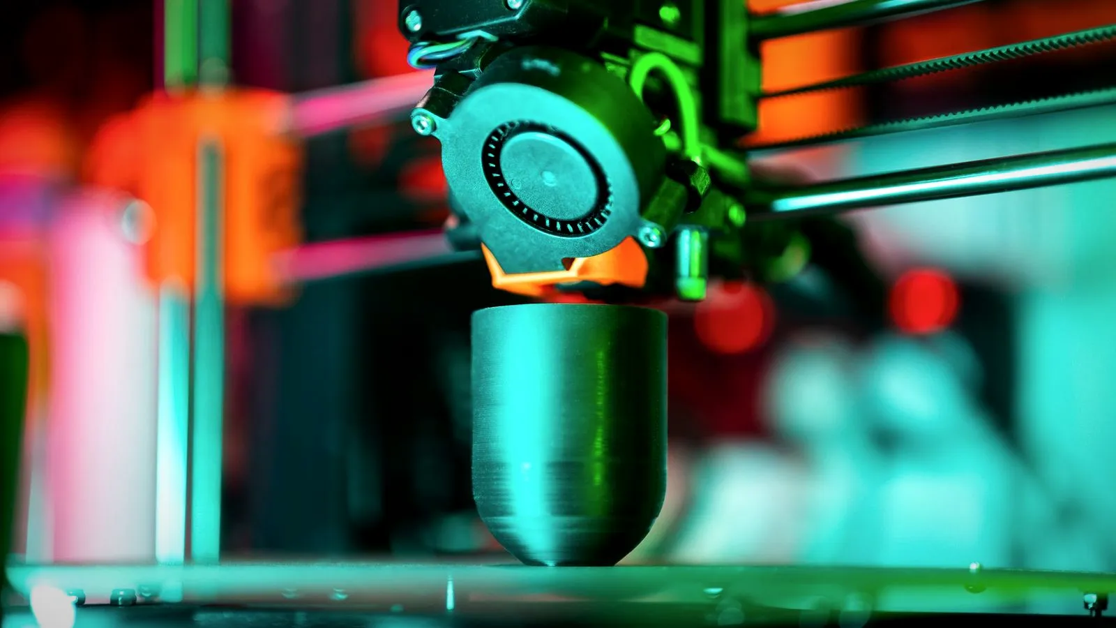 A 3D printer nozzle printing a rounded gray part in teal light