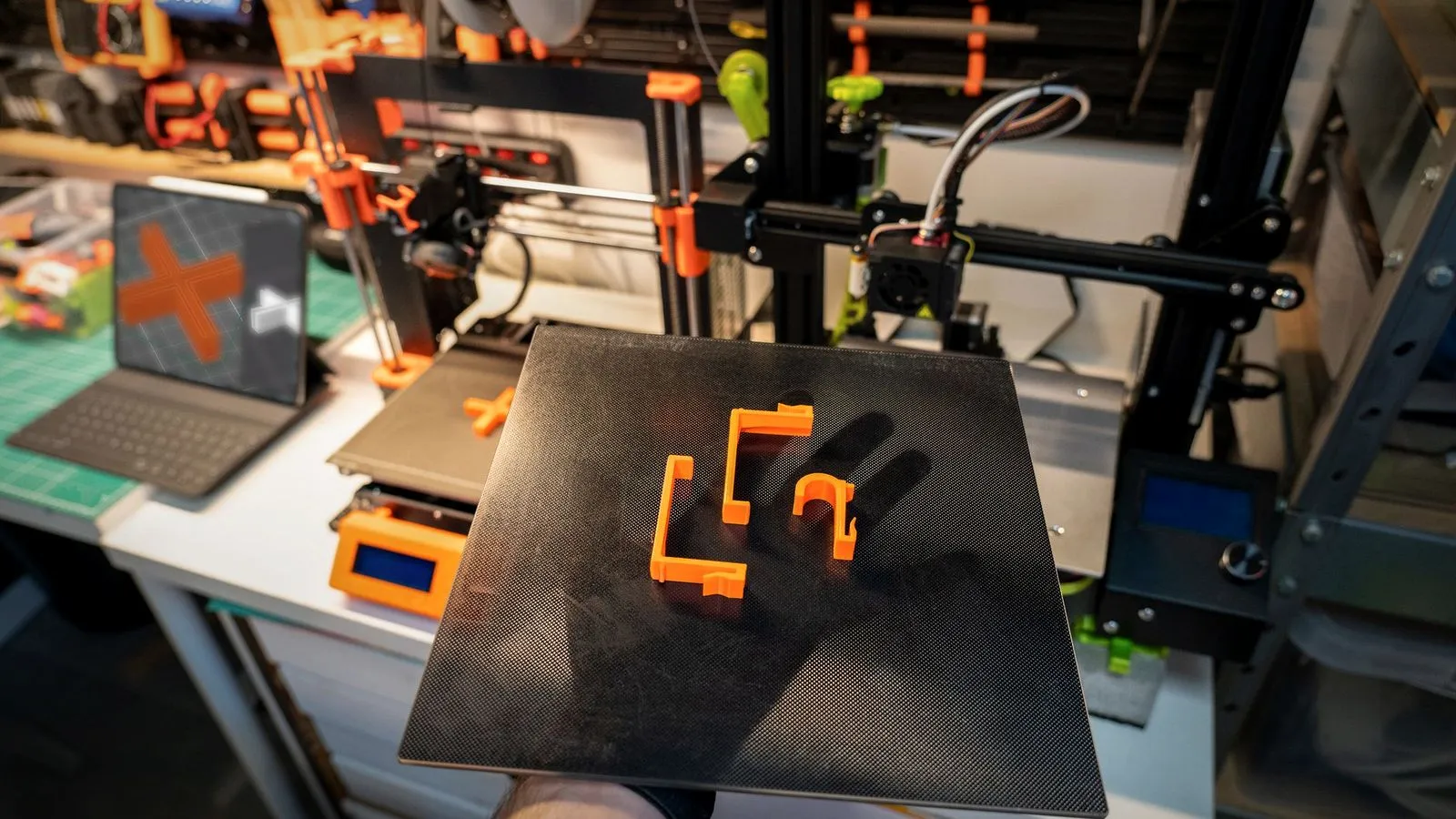 Hand holding a textured build plate with orange 3D printed clips and brackets in front of two printers