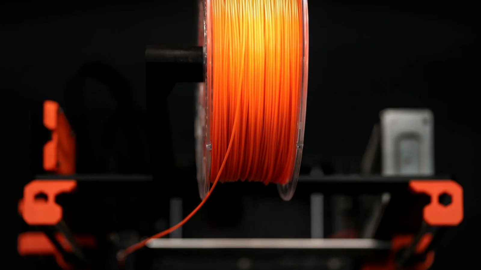 Spool of orange filament mounted on top of a 3D printer frame, with the strand running down toward the extruder
