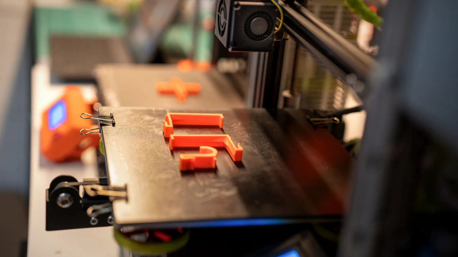 Orange printed parts on a black build plate fixed to the bed with binder clips