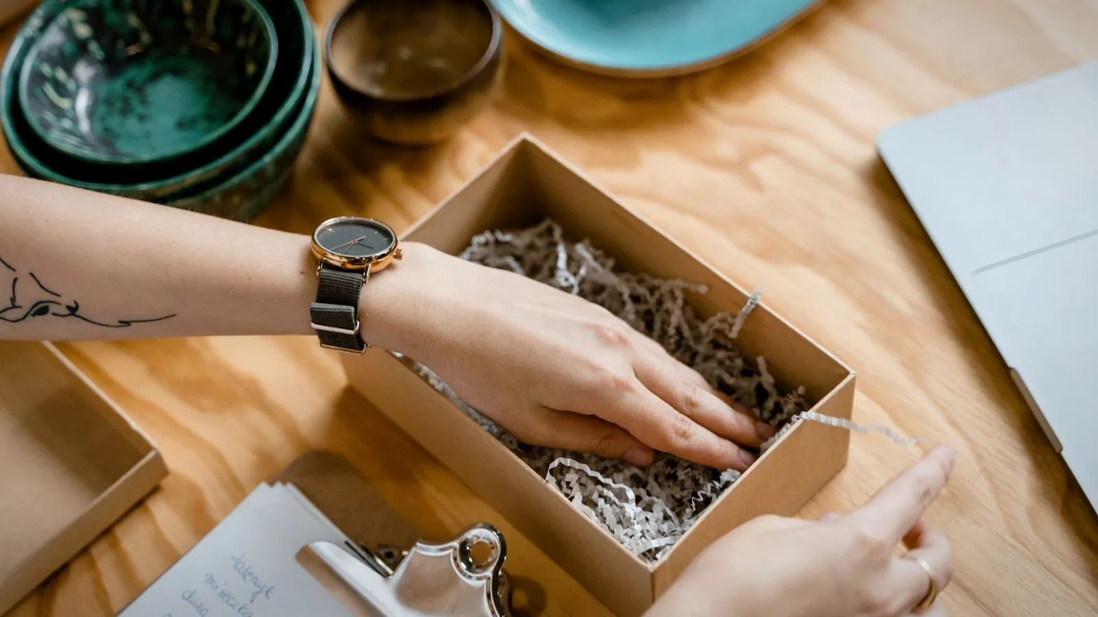 Hands packing a small cardboard box lined with shredded paper on a wooden desk