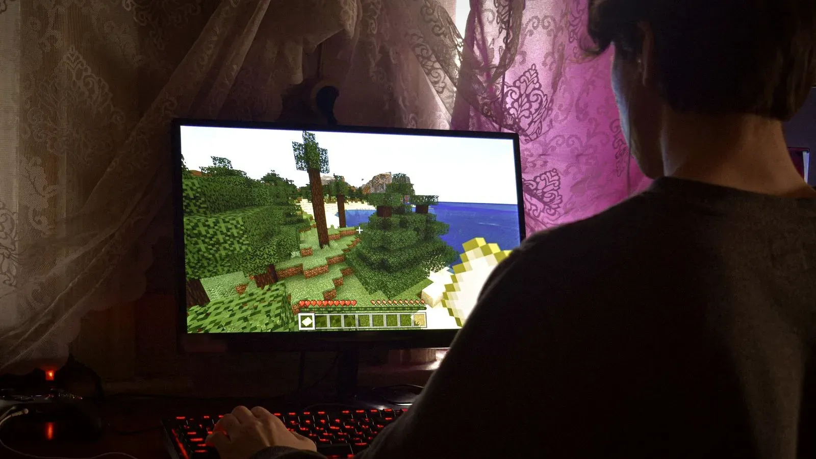 Person playing Minecraft on a desktop computer, with a blocky forest landscape on the monitor