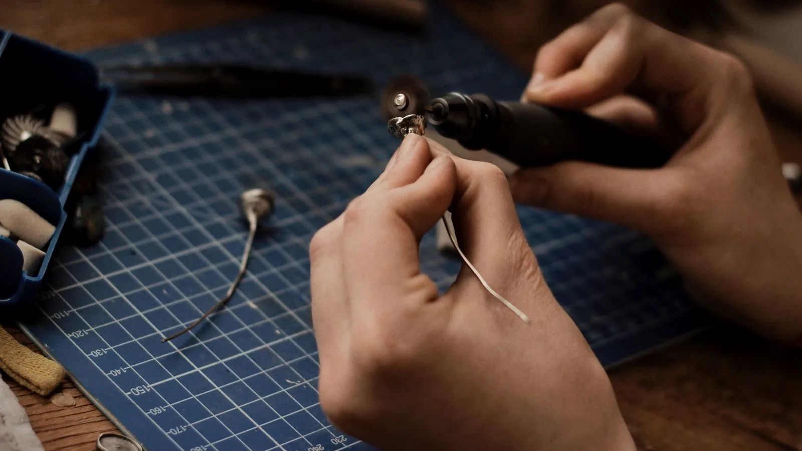 Hands polishing a small silver piece with a rotary tool over a cutting mat