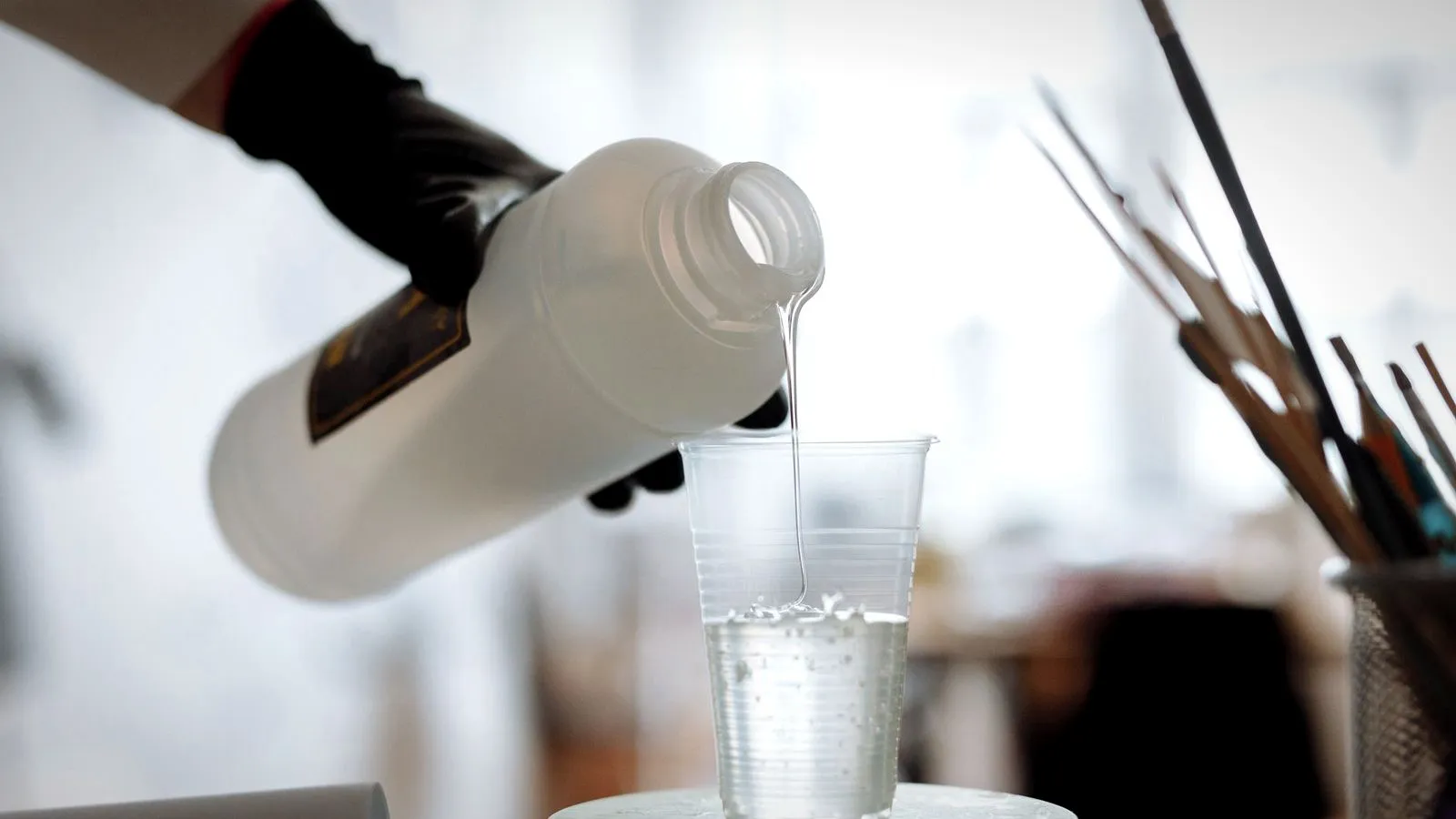 Gloved hand pouring a clear liquid from a bottle into a plastic cup on a scale
