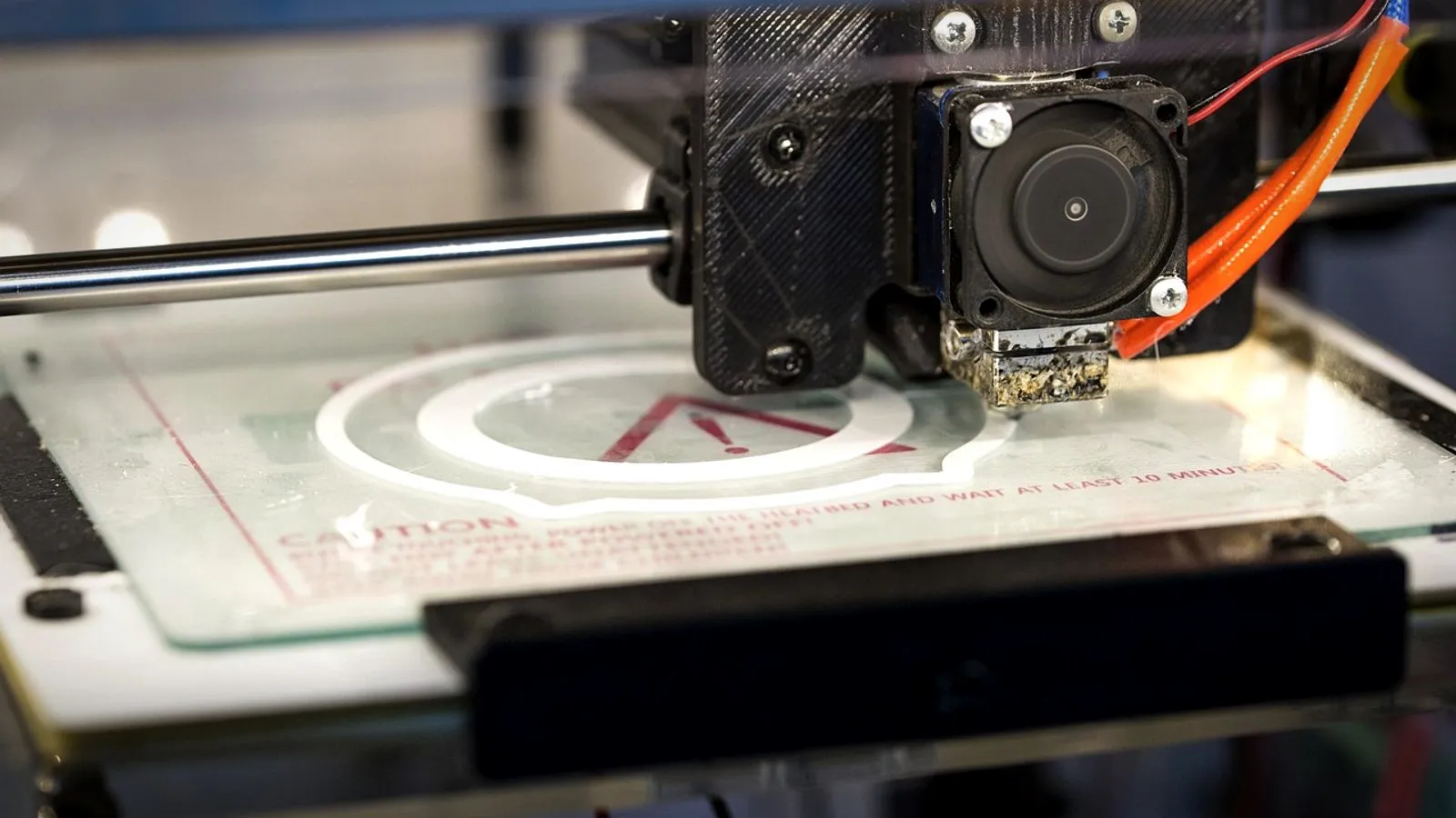 3D printer print head laying the white first layer of a round part on a glass bed, with plastic residue on the nozzle