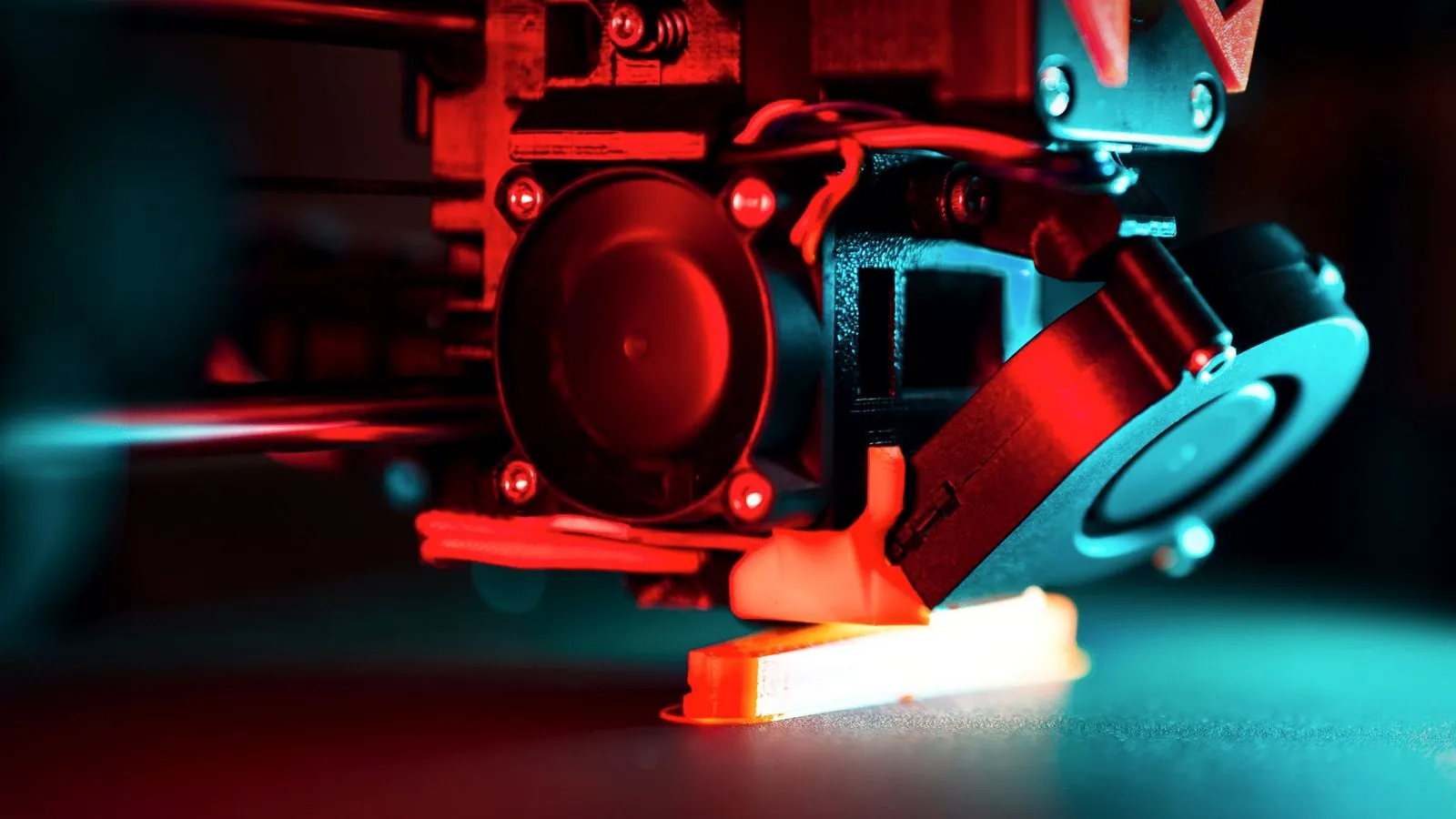 3D printer print head laying down an orange part under red and teal light
