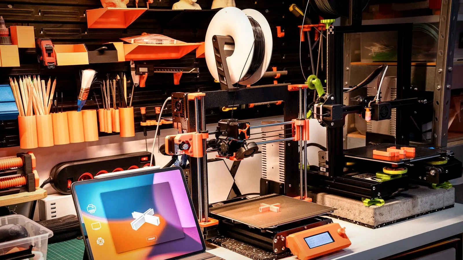 A small print workshop bench with two desktop 3D printers, a tablet showing a model and printed parts on the shelves