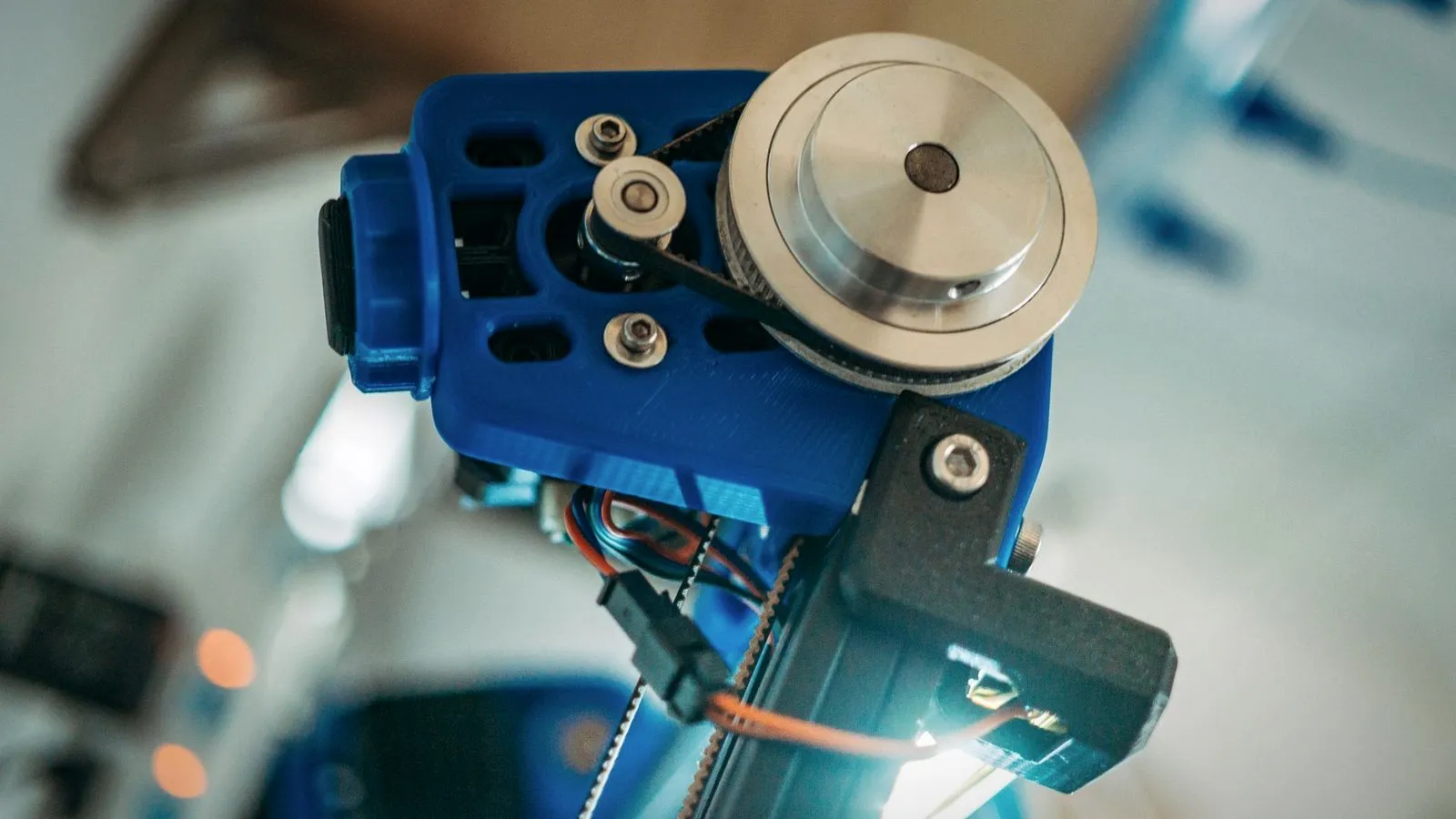 Blue 3D printed mount carrying an aluminum pulley and toothed belt, with a black printed bracket below