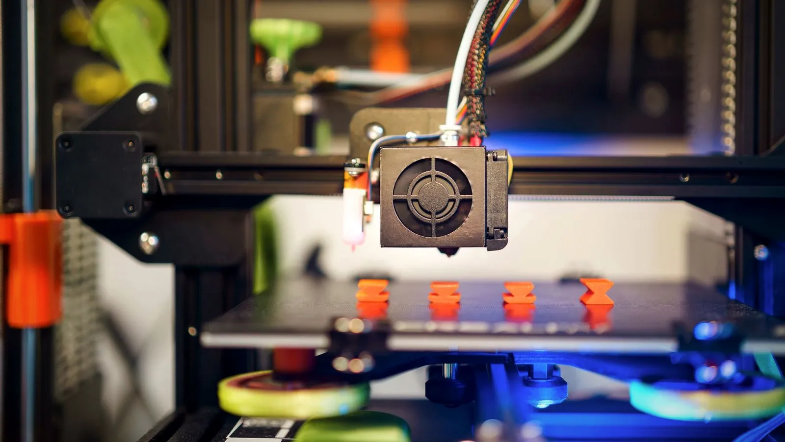 Print head of a 3D printer above four small orange test prints, with yellow bed leveling knobs below the bed