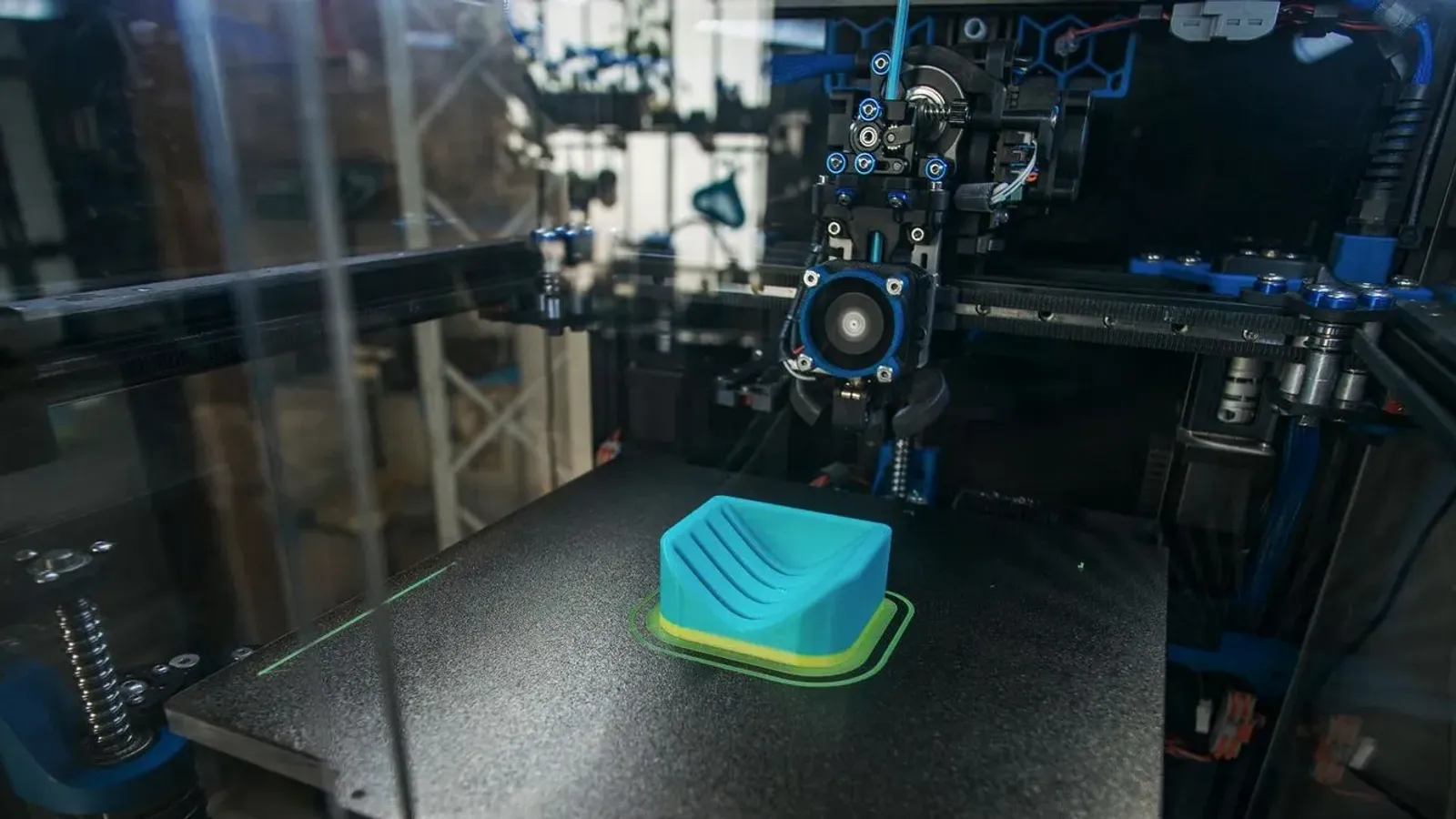 Teal part printing on a textured build plate inside a printer enclosed by clear panels