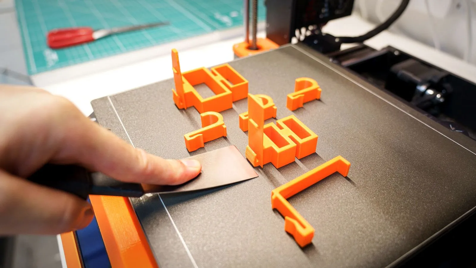 Hand sliding a thin spatula under orange 3D printed parts on a black build plate