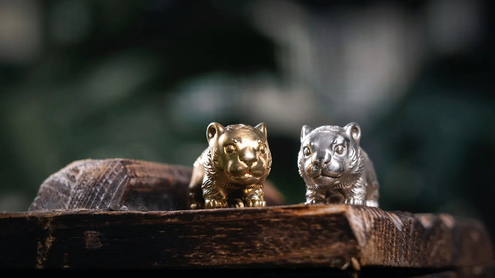 Two small metal tiger figurines, one brass-colored and one silver, on a rough wooden board