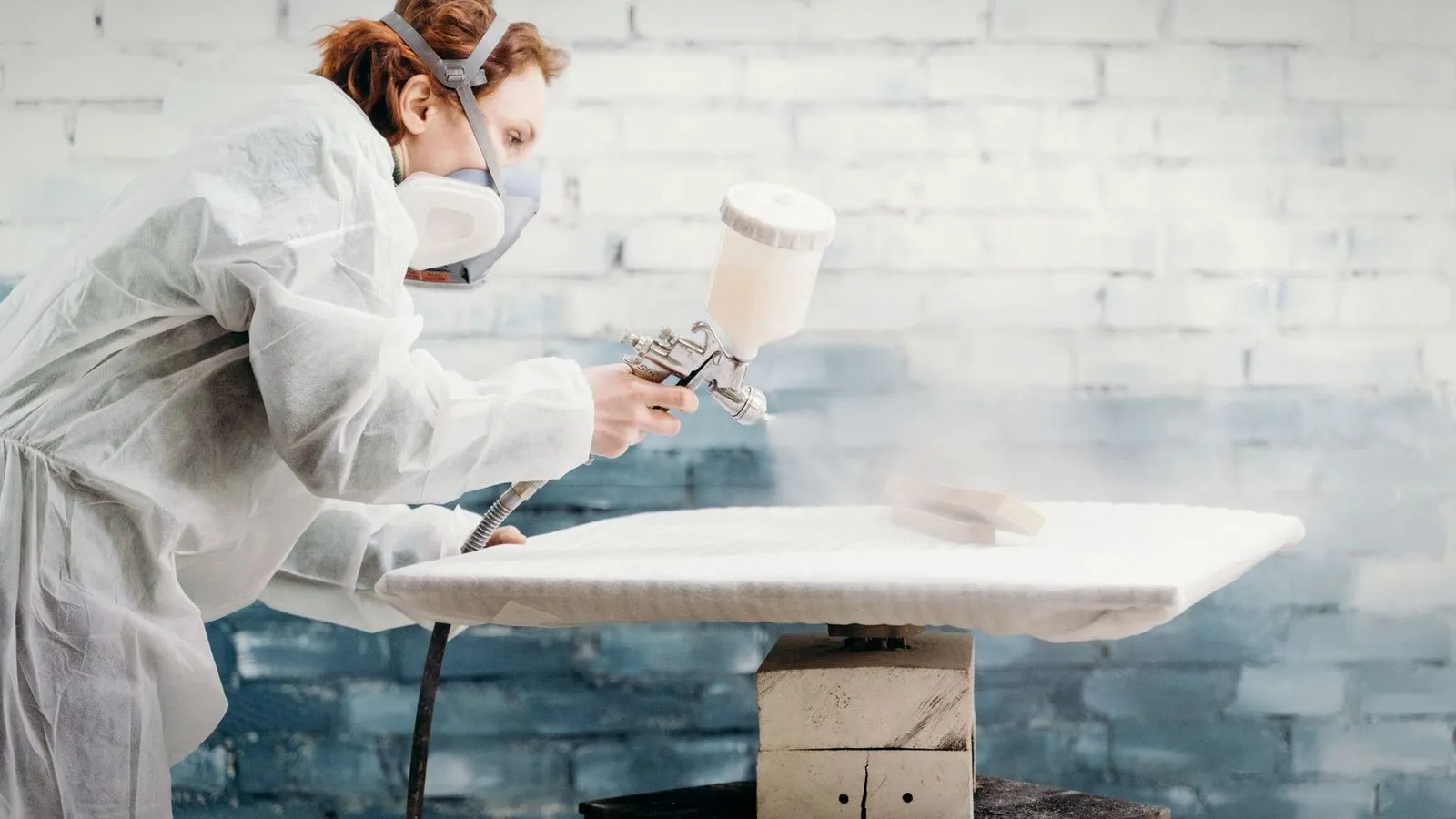 Woman in a protective suit and respirator spraying paint onto a panel with a spray gun