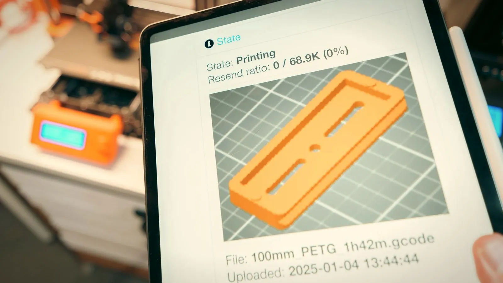 Tablet screen showing a print job’s state and a preview of the part, with the 3D printer blurred behind it