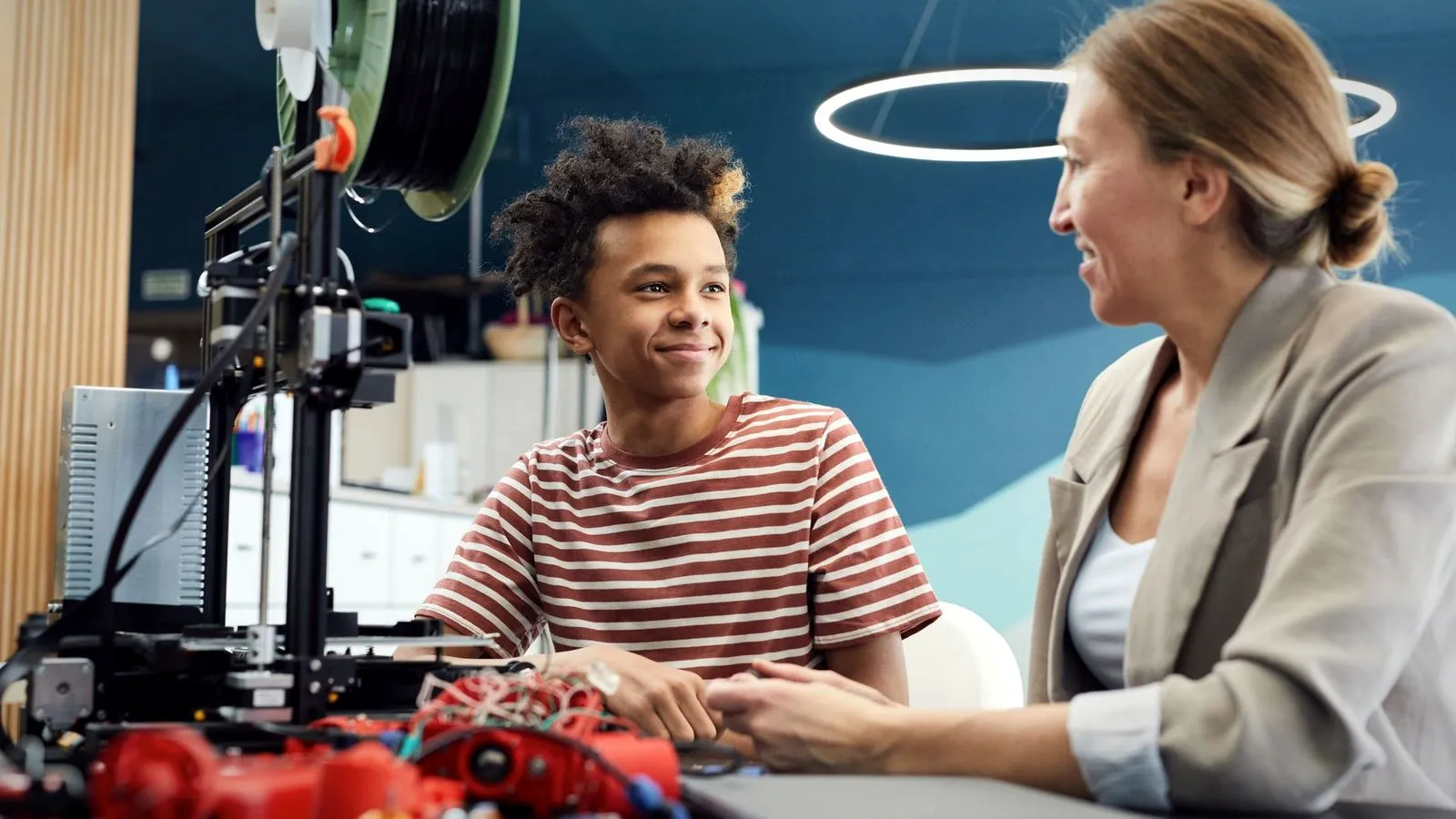 A teenager and an adult talking at a table next to a 3D printer and red printed parts