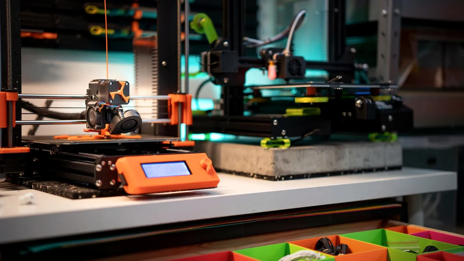 Two 3D printers side by side on a workbench above an open drawer of colorful printed bins