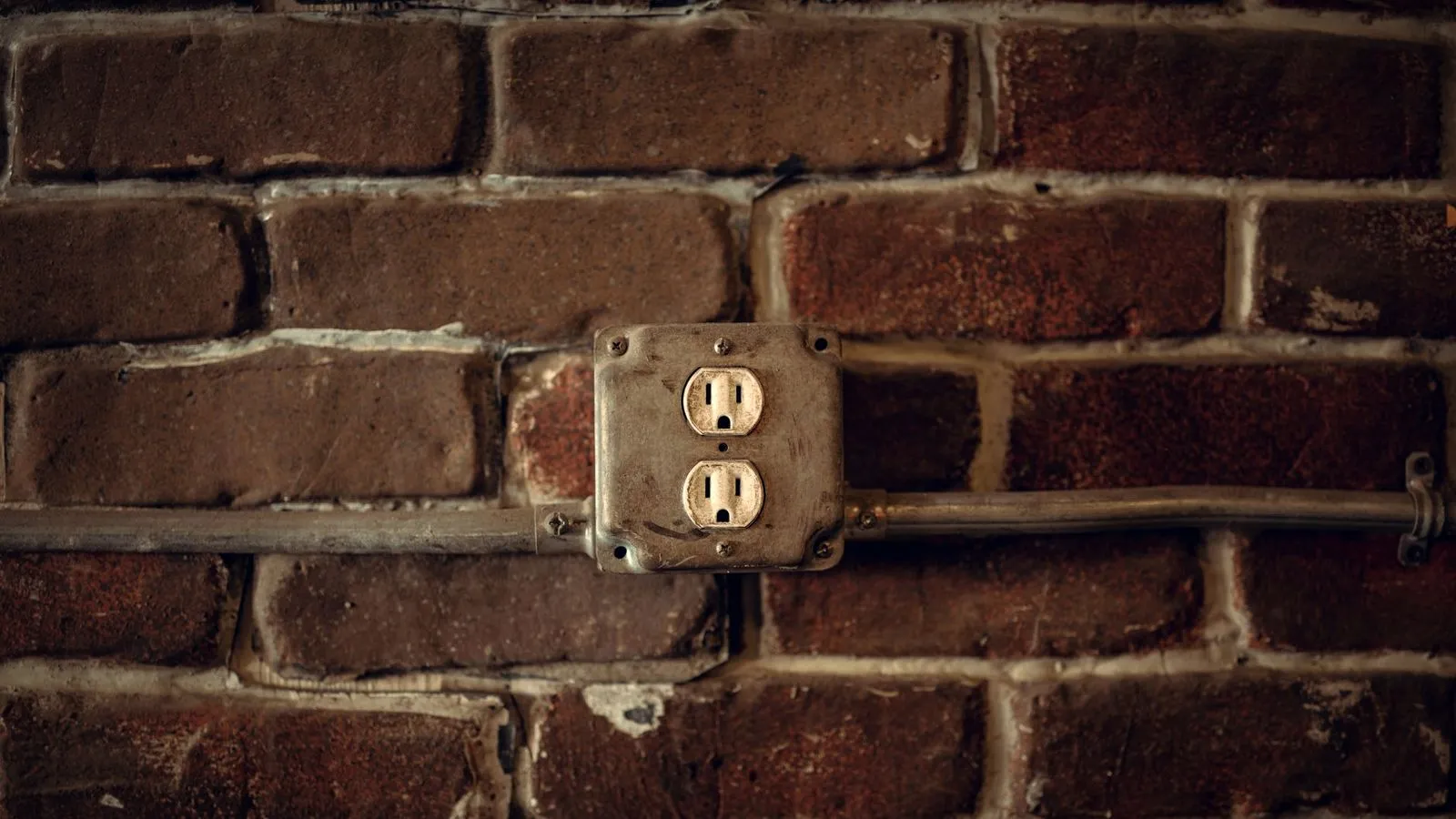 A duplex wall outlet in a metal box with conduit running along a red brick wall